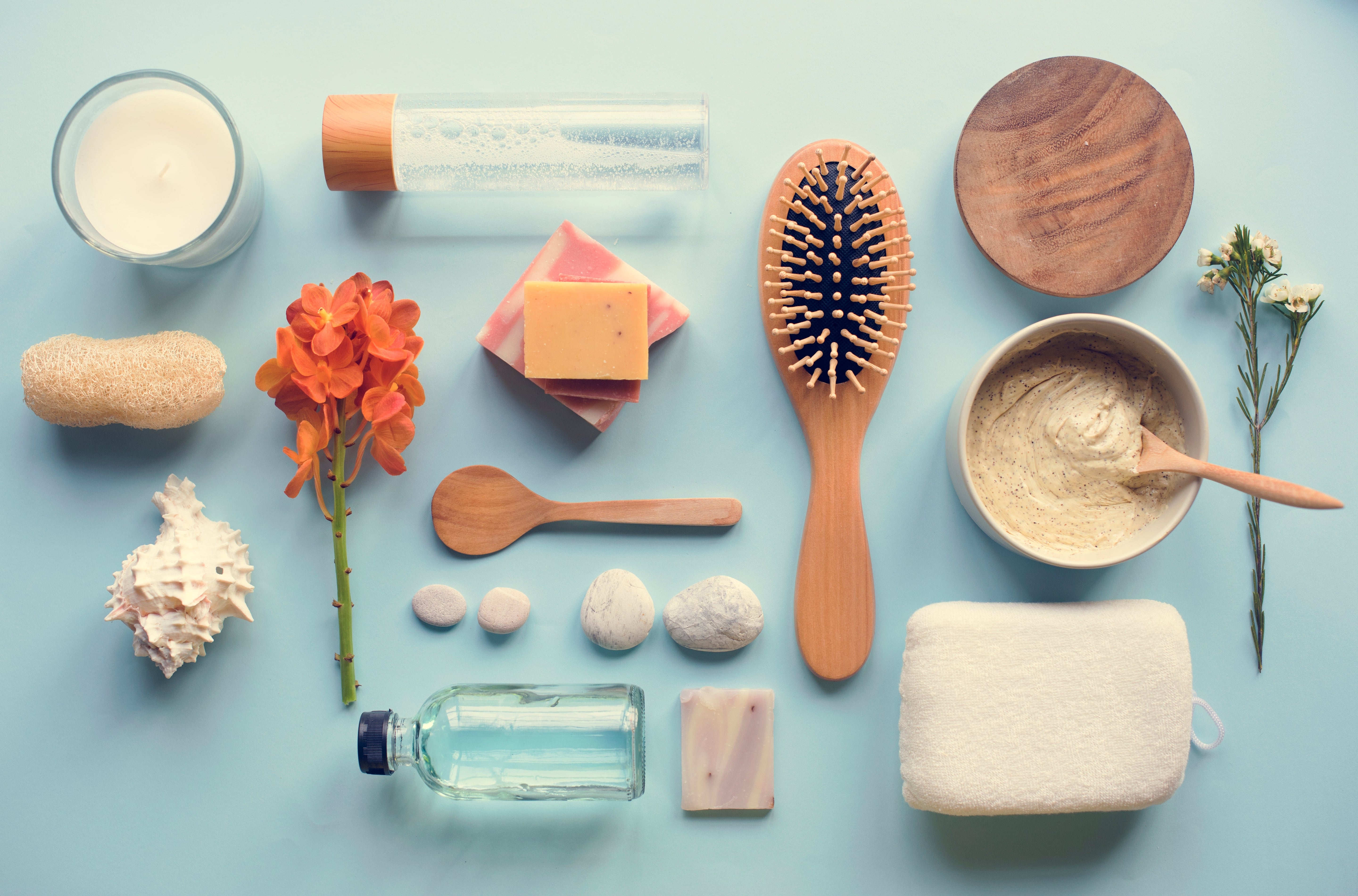 A flat-lay of home essentials on a pastel blue background, featuring a hairbrush, soaps, a candle, a loofah, a towel, a glass bottle, a wooden spoon, and fresh flowers.