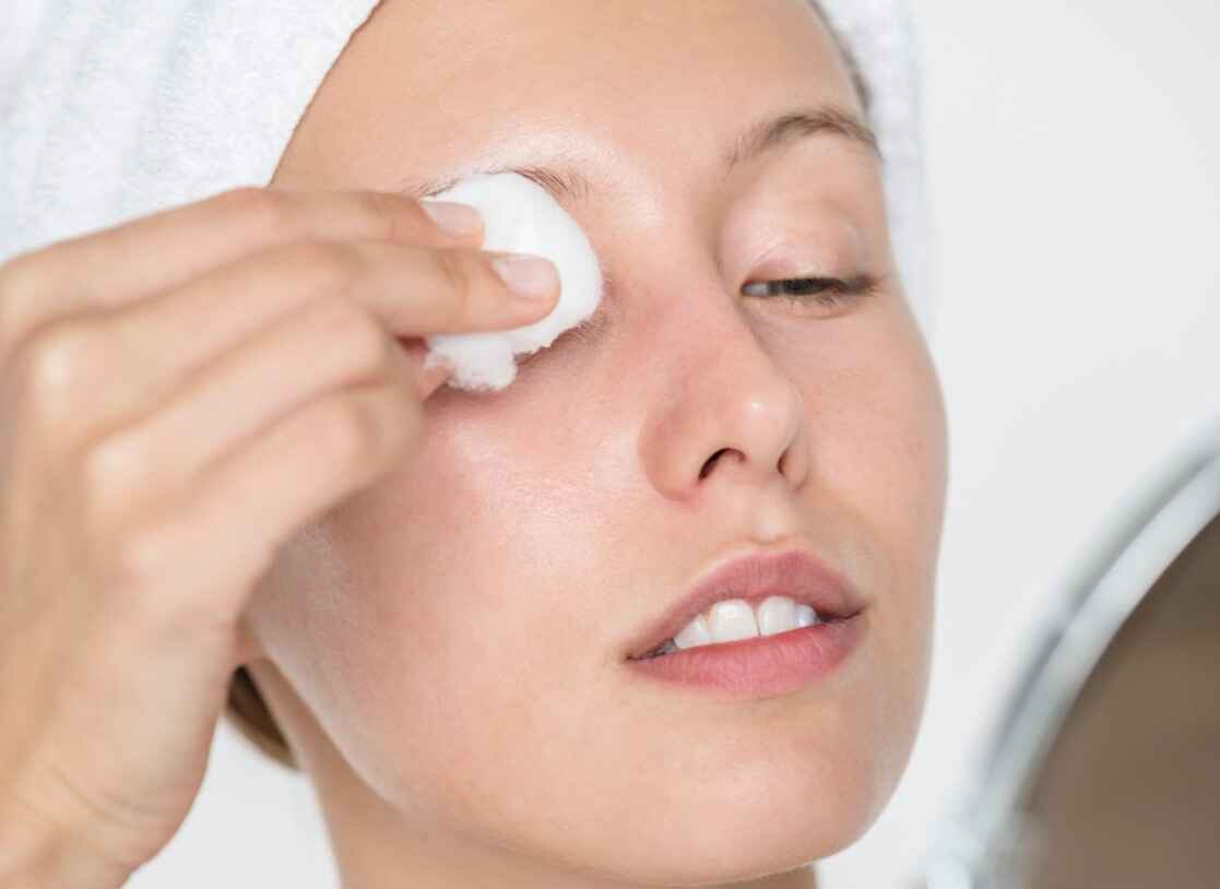 A close-up of a woman with a white towel wrapped around her head, gently applying an exfoliating gel to her face using a cotton pad. 