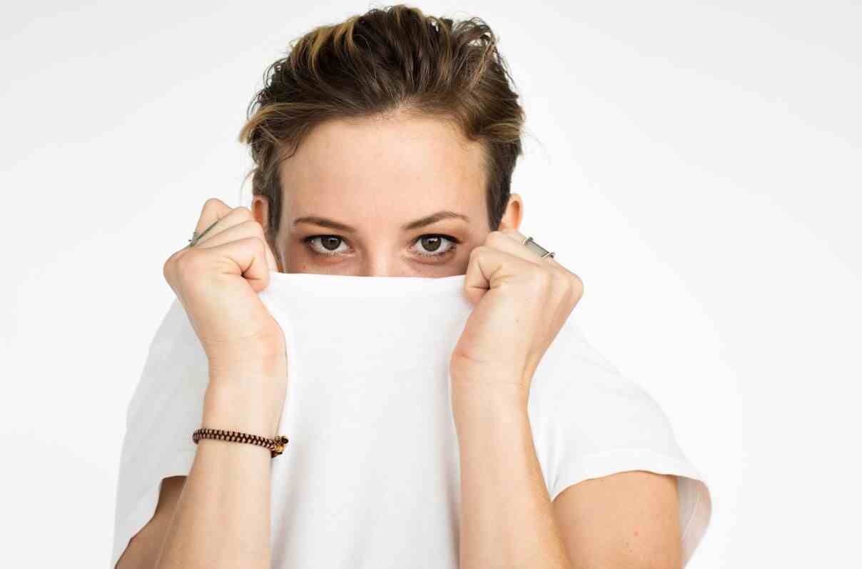 A woman with under eye patches gazes directly at the camera, partially covering her face with a white shirt. Her eyes are expressive, and the patches help reduce puffiness.